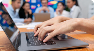Hands typing on a laptop during a professional meeting with a team in the background.