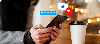A person's hands holding a black smartphone at a wooden table with a white coffee cup nearby, illuminated by warm bokeh lights, with floating social media icons (stars, blue chat bubble, blue like thumb, red heart, yellow lightning bolt) above the screen.
