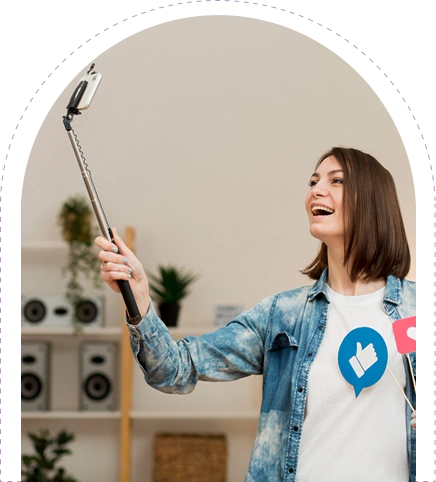 A smiling young woman taking a selfie with a selfie stick, wearing a white t-shirt with a large blue Facebook "Like" thumb icon and a speech bubble.